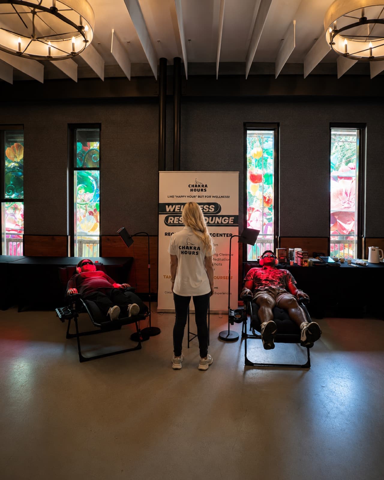Two guests reclining in zero-gravity chairs under red light with a facilitator between them at a Chakra Hours Wellness Reset Lounge in a Dallas venue, Chakra Hours