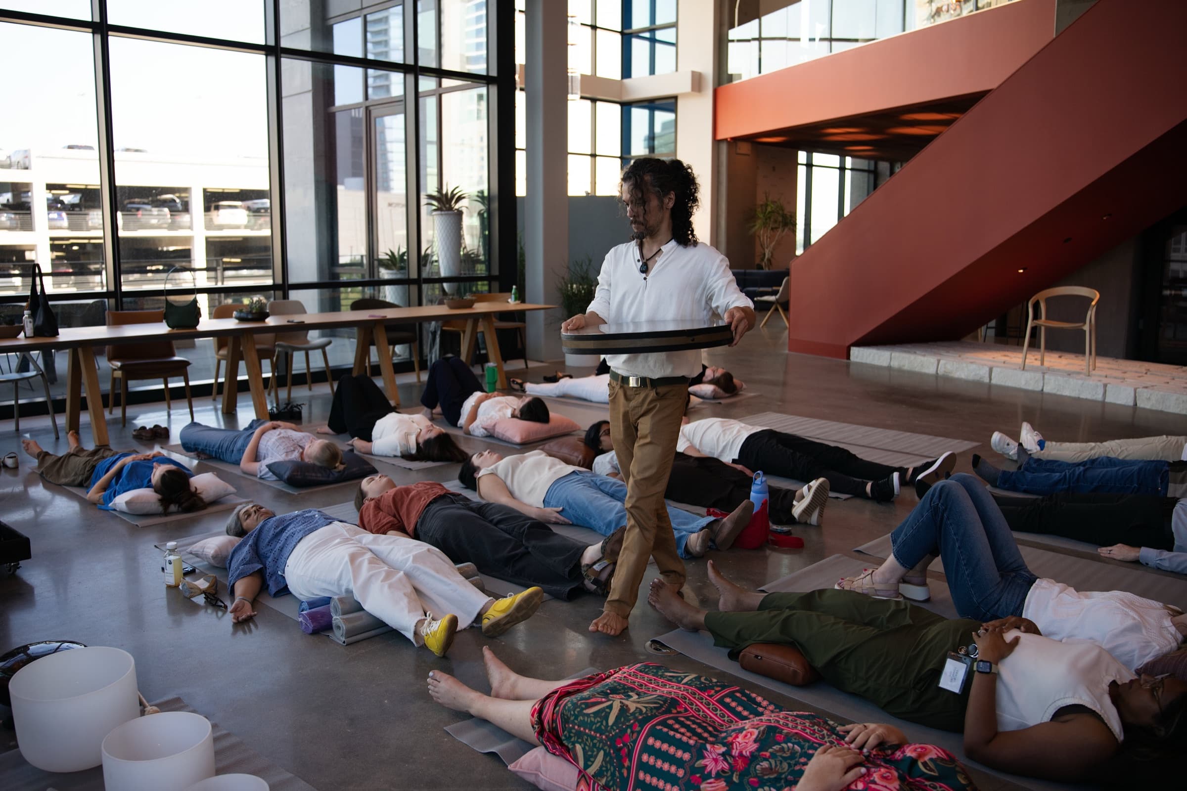 Chakra Hours facilitator carrying an ocean drum over employees resting on mats during a live corporate sound bath in a Dallas office